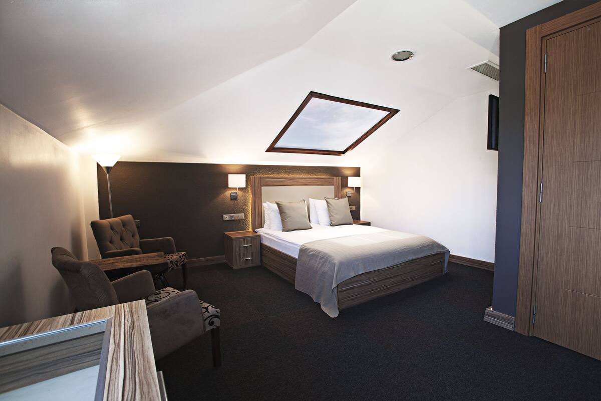 A hotel room with a bed, a desk, a chair, a nightstand, and a wooden wardrobe.