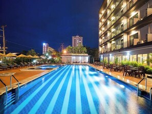 A swimming pool with lounge chairs and tables, surrounded by a building with balconies and a view of a tall building in the background. - Savotel (Sattahip)