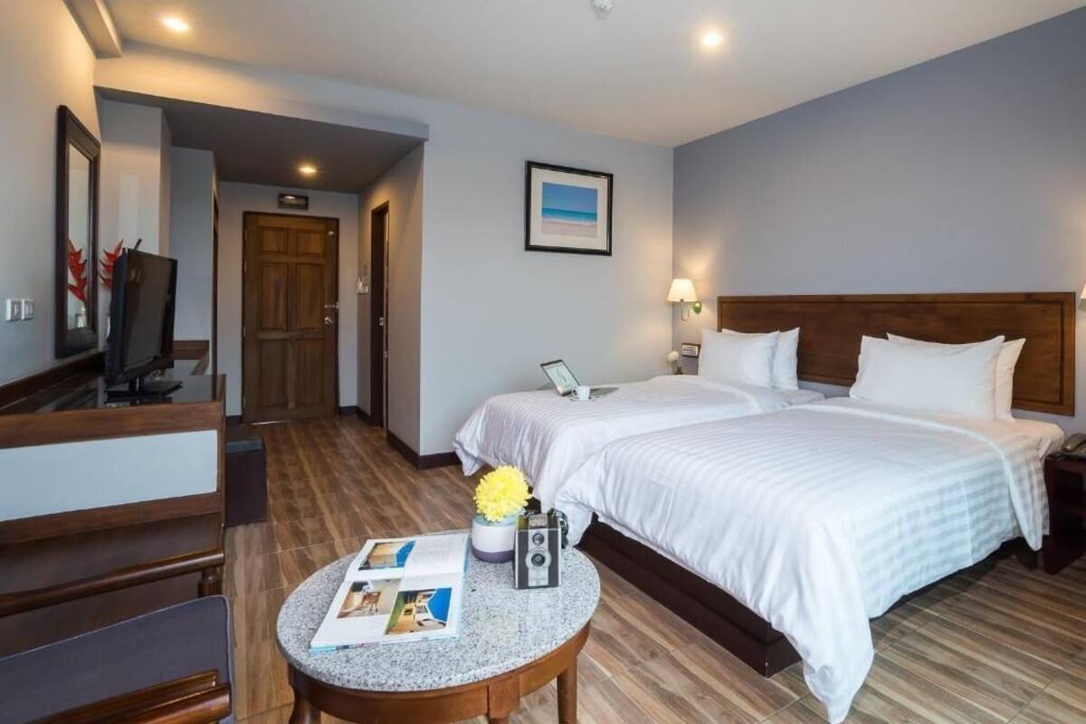 A hotel room with two beds, a desk with a chair, a small table with a lamp, and a framed picture on the wall.