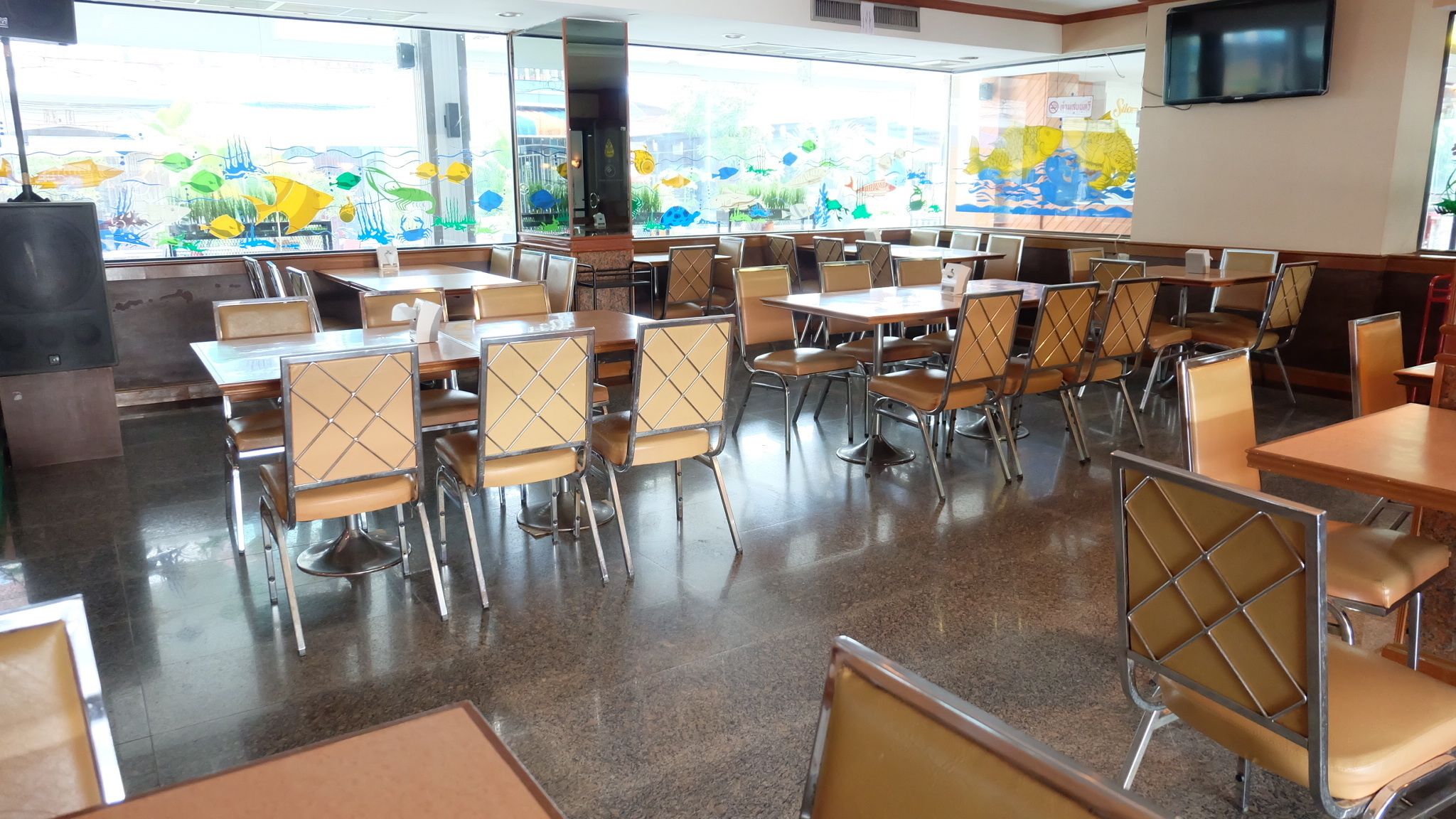 A dining area with tables and chairs, large windows with artwork, and a flat-screen TV on the wall.