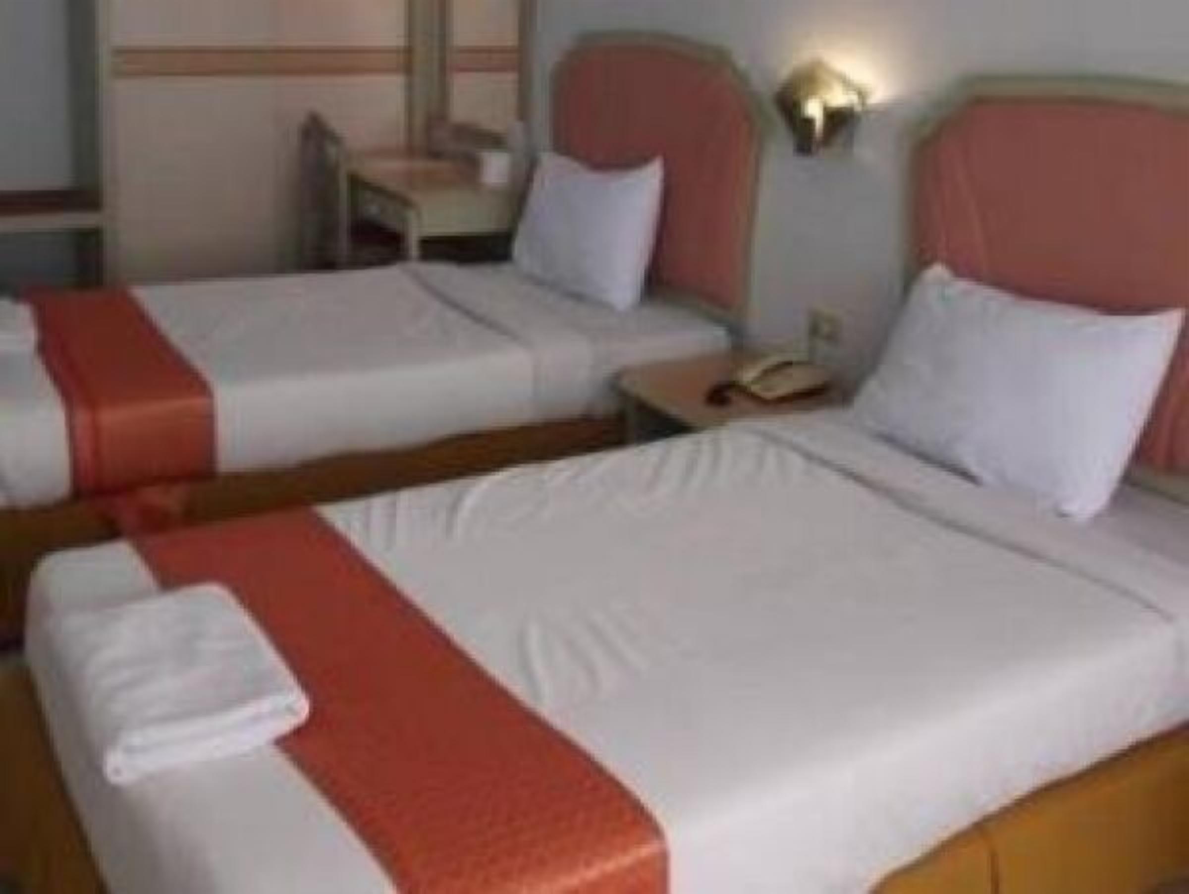 Dos camas individuales con sábanas blancas y detalles rojos, una mesita de noche con lámpara, y una silla pequeña en una habitación de hotel.
