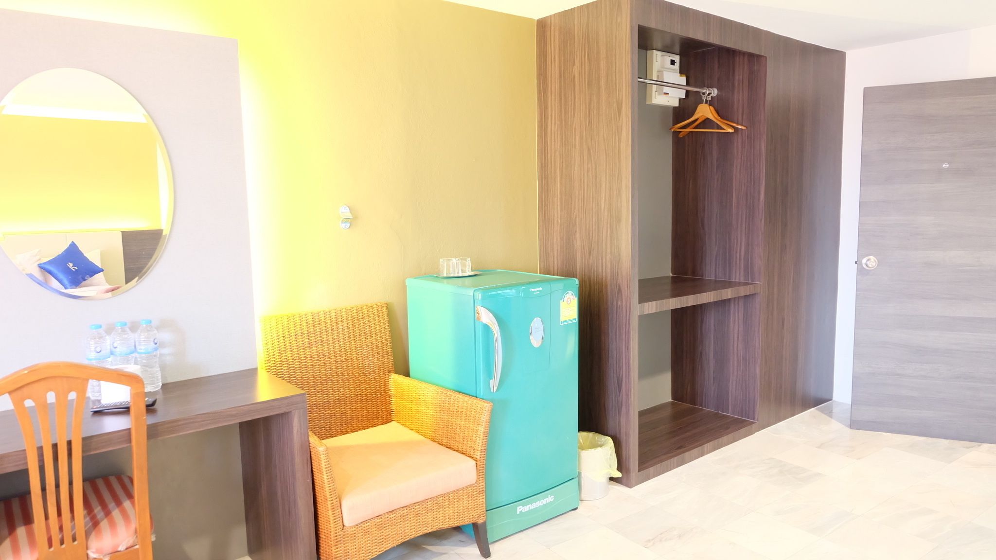 A compact room with a teal refrigerator, wicker chair, and a wooden wardrobe with a hanging rack.