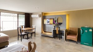 A hotel room with a bed, a dining table with chairs, a sitting area with a chair, a small table with a vase, a wall-mounted mirror, and a green refrigerator. - Silom Boutique Hotel Hatyai (Hat Yai)
