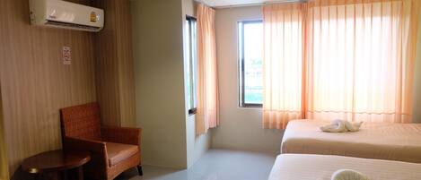 A hotel room with a single wicker chair, two beds, a window with curtains, and an air conditioning unit.