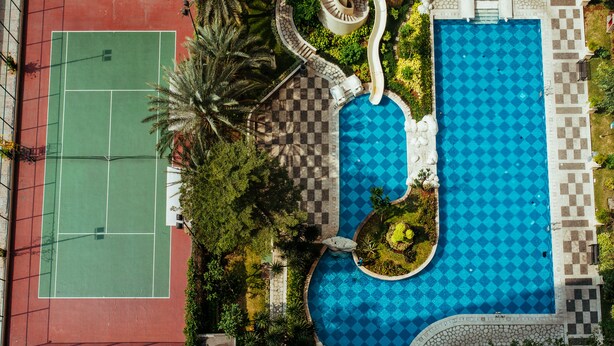 An aerial view of a resort with a tennis court, a swimming pool with a checkered pattern, and surrounding landscaped areas.