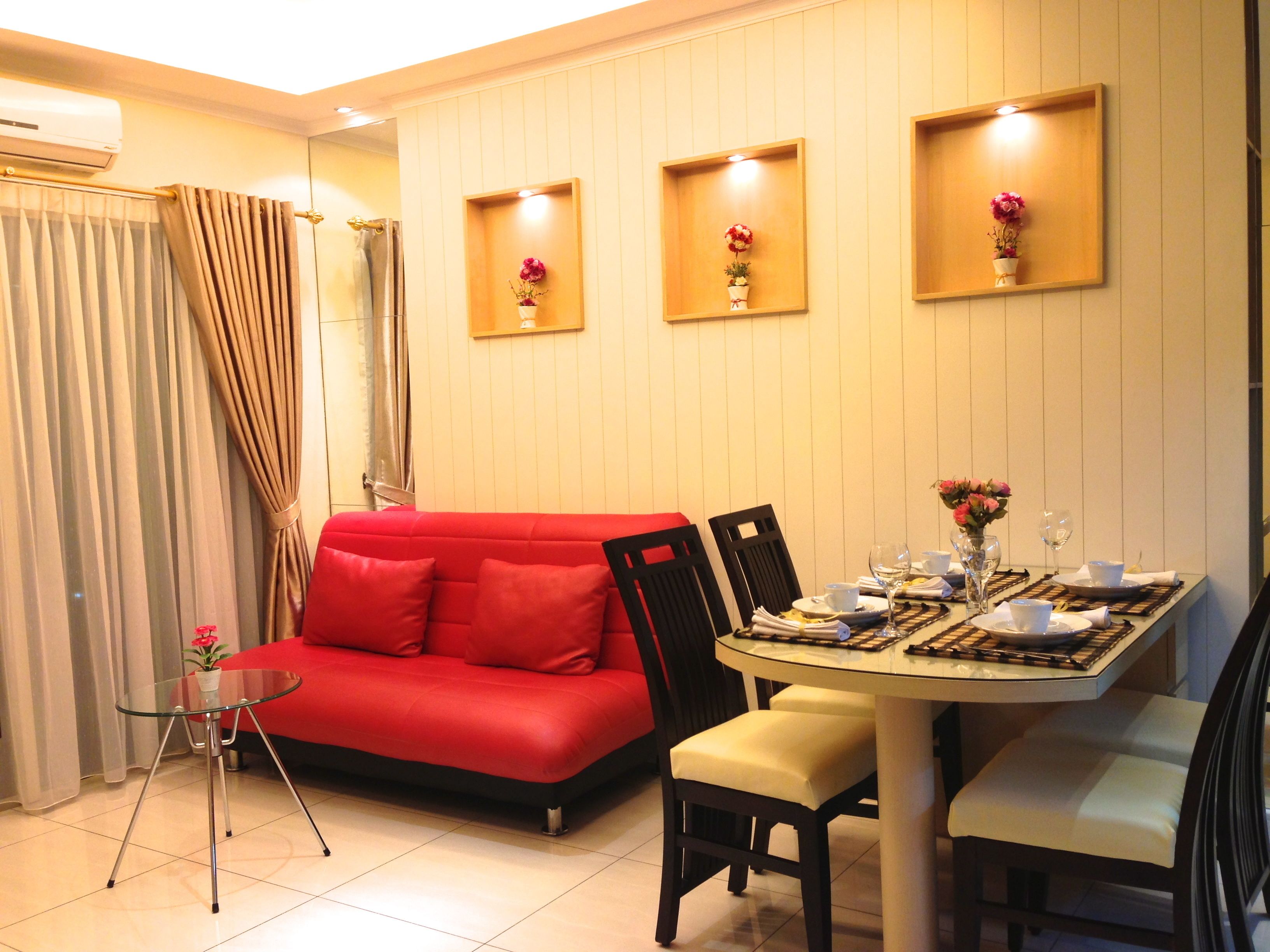 A dining area with a glass table, chairs, and a red sofa. There are three wall-mounted shelves with flowers.