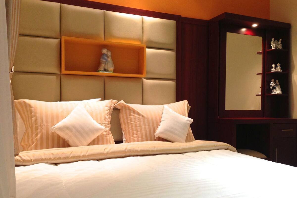 Quarto de hotel com uma cama grande, cabeceira com prateleira embutida e armário de madeira com objetos decorativos.