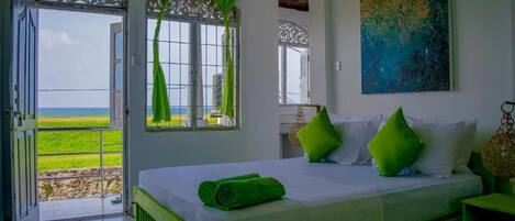 A bedroom with a bed, green pillows, a view of the sea, and a painting on the wall.