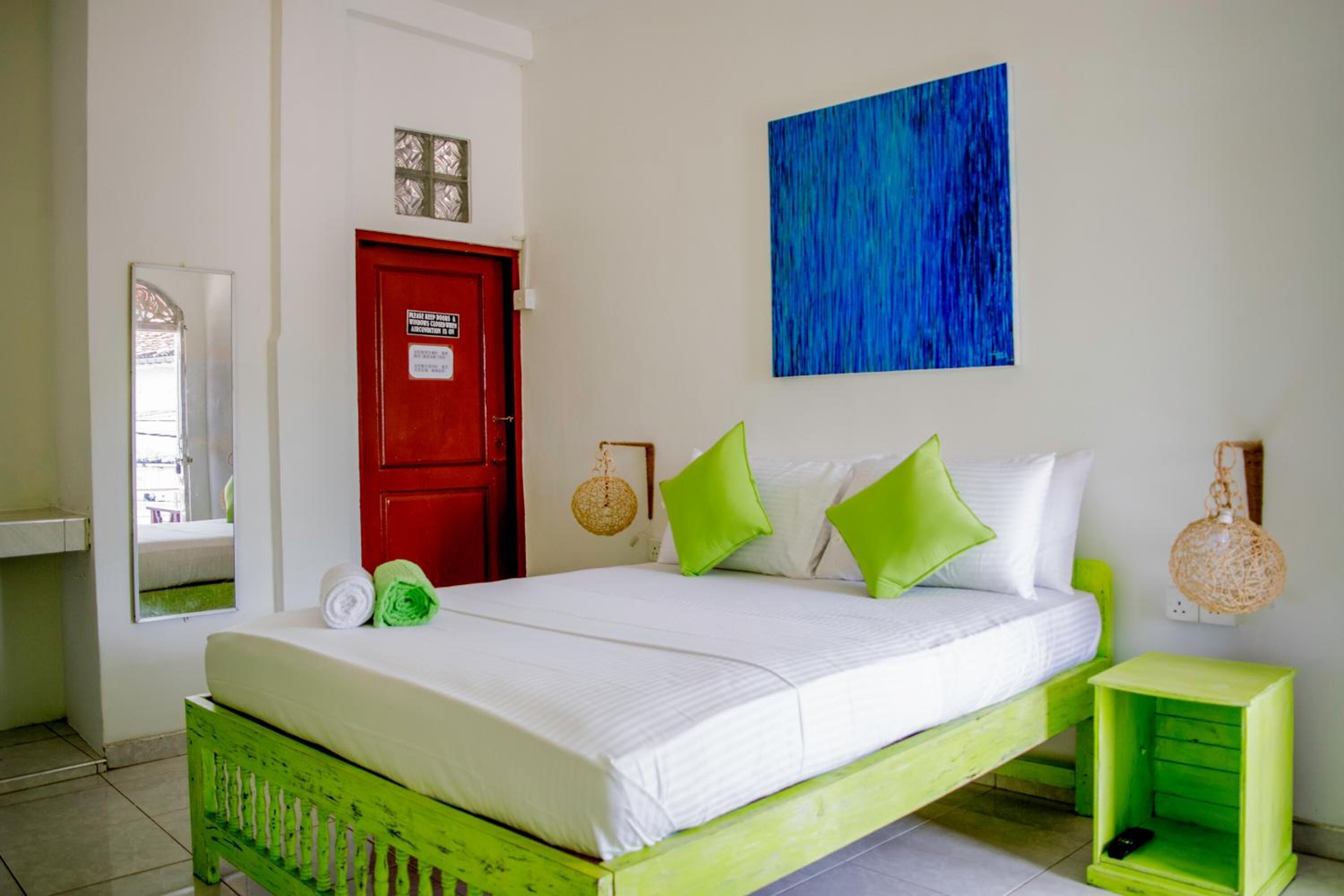 A bed with green and white bedding, a red door, a blue abstract painting, and a small green nightstand with a phone.