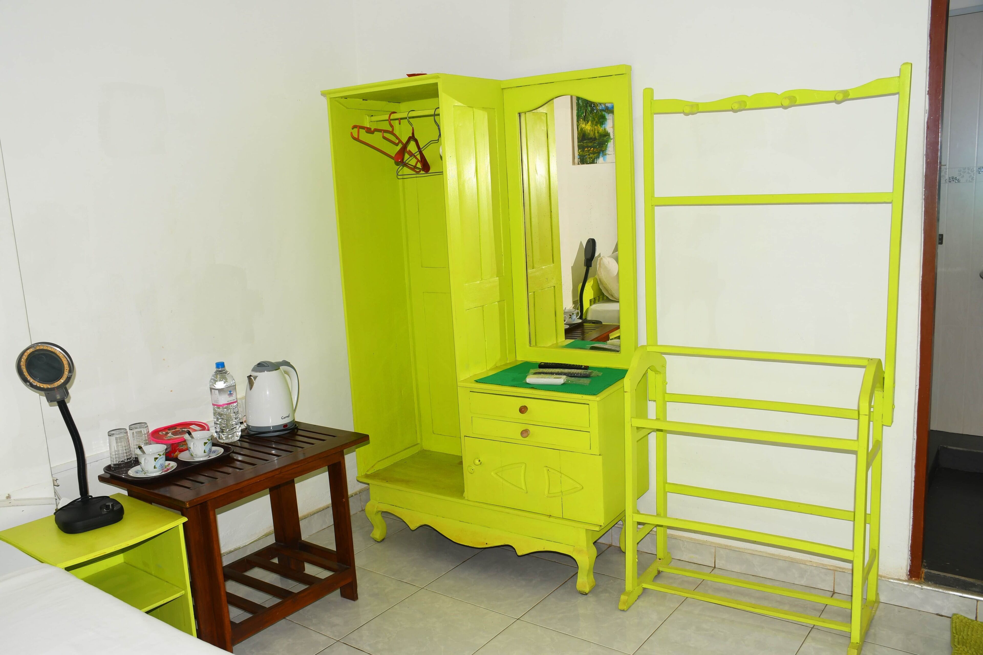 A small, brightly colored room with a green vanity, a mirror, a small table with a kettle, cups, and a water bottle.