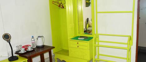 Una stanza piccola e luminosa, con un comò verde, uno specchio, un tavolino con un bollitore, tazze e una bottiglia d'acqua.