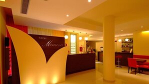 Zona de recepción de hotel con diseño moderno, que incluye un mostrador de recepción, un área de estar con sillas rojas y un bar con estantes llenos de botellas.