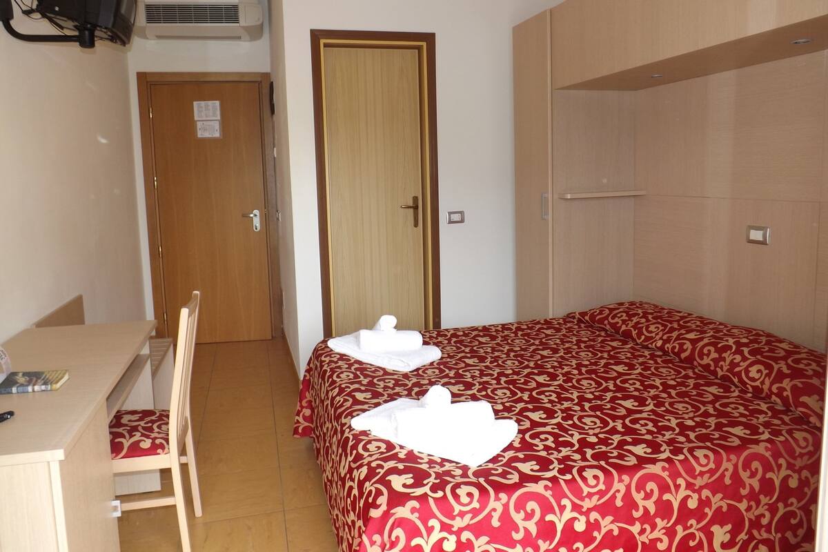 A hotel room with a bed, a desk, and a chair. There is a door and an air conditioning unit on the wall.