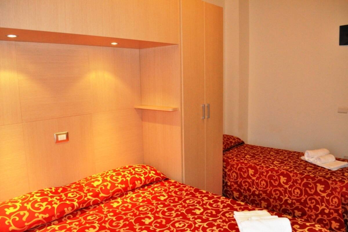 A hotel room with two beds, each with a red patterned bedspread and white towels. There is a wooden wardrobe with a shelf and a mirror.