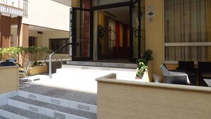 Entrada de hotel con puerta de vidrio, escalones que la conducen y mobiliario exterior.