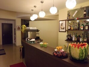 Uma área de bar com uma máquina de café, uma exposição de frutas e uma prateleira com garrafas de bebidas.
