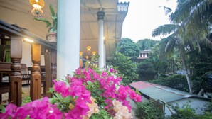 A covered outdoor area with wooden furniture, a potted plant, and a view of a building with a red roof. - La Villa Guest House (Unawatuna)