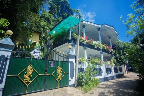 Un bâtiment avec une porte verte, une enseigne indiquant « La Villa », et un balcon orné de plantes en suspension.