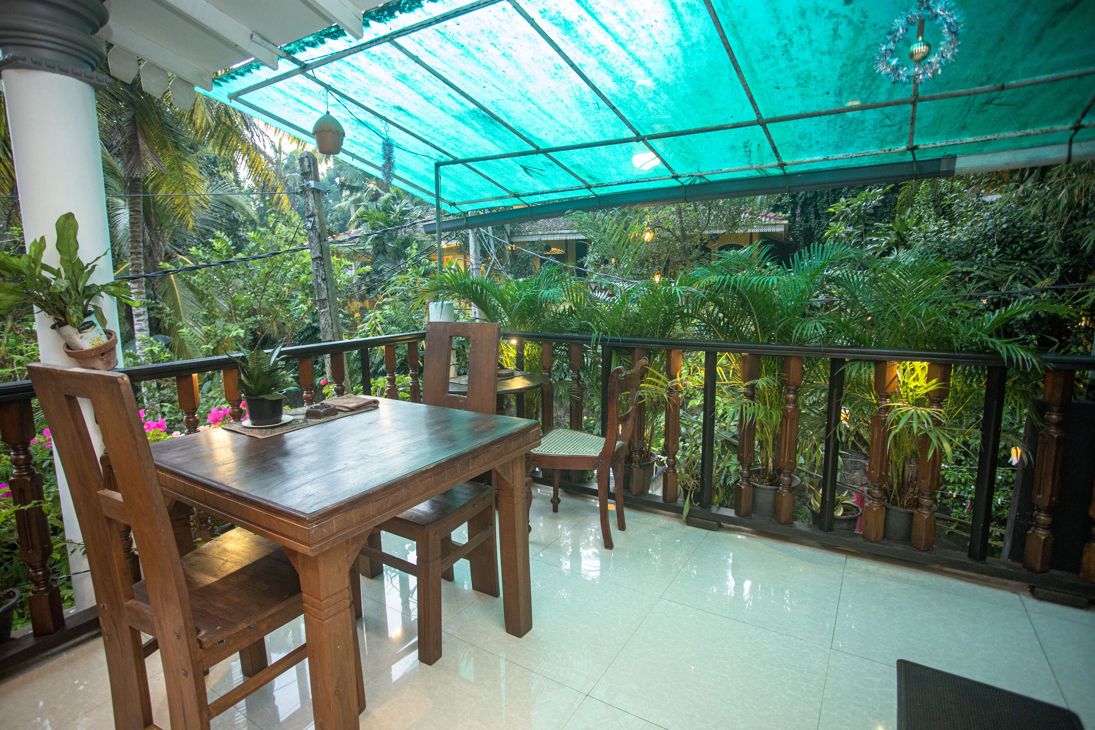 Un área de comedor al aire libre techada, con una mesa y sillas de madera, rodeada de plantas en macetas y vistas a follaje exuberante.