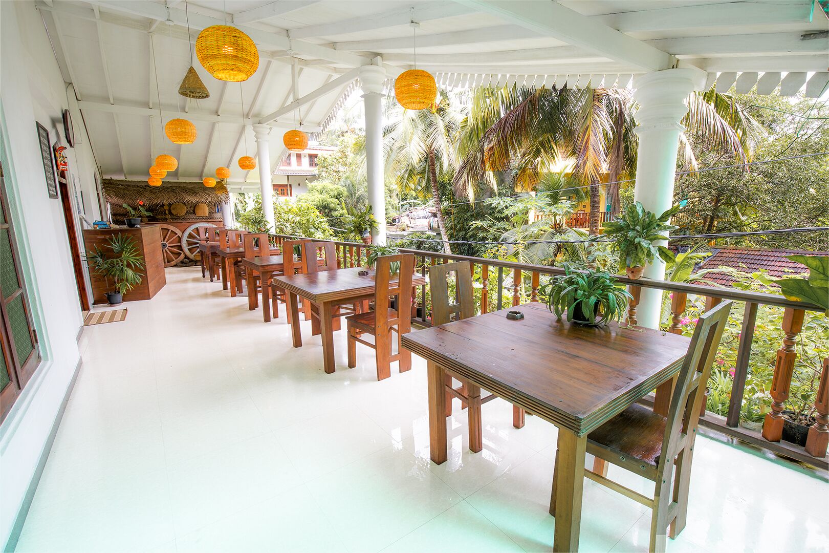 A covered outdoor dining area with wooden tables and chairs, potted plants, and hanging lanterns.