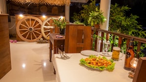 An outdoor dining area with a table set for two, featuring a salad, a beer mug, and a bottle. There are wooden chairs, a thatched roof, and potted plants in the background. - La Villa Guest House (Unawatuna)