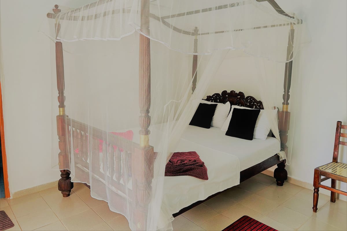 Een bed met hemelbed voorzien van wit beddengoed en zwarte kussens, een houten stoel en een rood vloerkleed op een betegelde vloer.