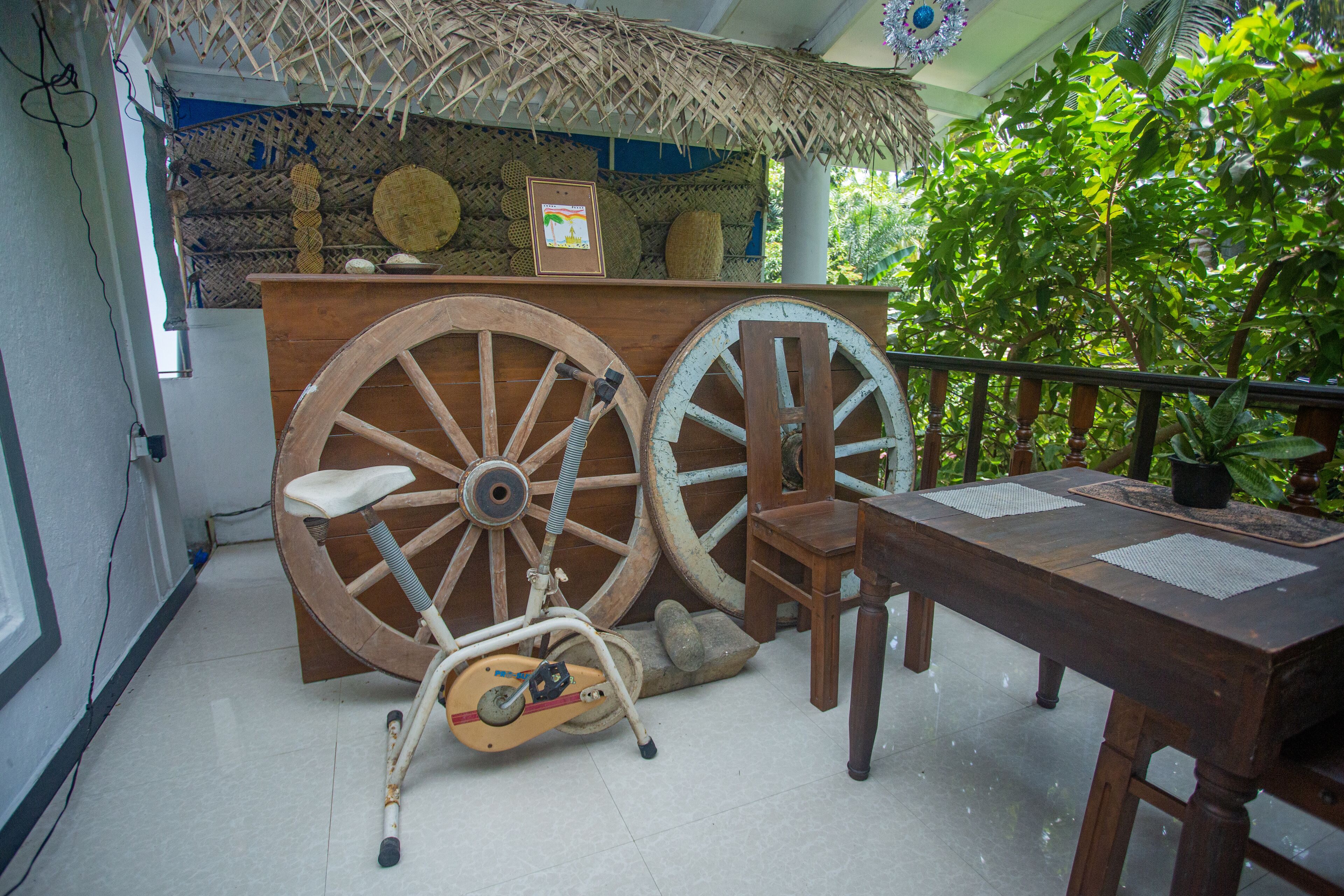 Une table en bois avec une chaise, un vélo et une grande structure en forme de roue, dans une pièce dont le plafond est recouvert de chaume et où l’on trouve des plantes vertes.