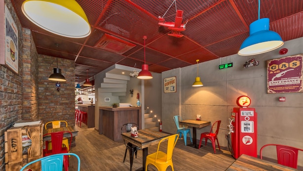 Interior de restaurante con paredes de ladrillo, mesas de madera, sillas coloridas y detalles rojos en el techo.