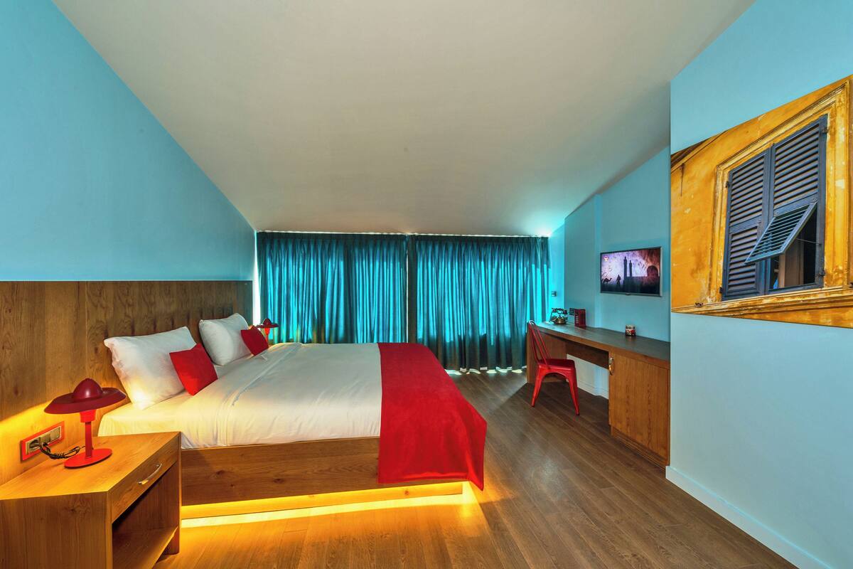 A hotel room with a bed, wooden headboard, a desk, a chair, and a television on the wall.