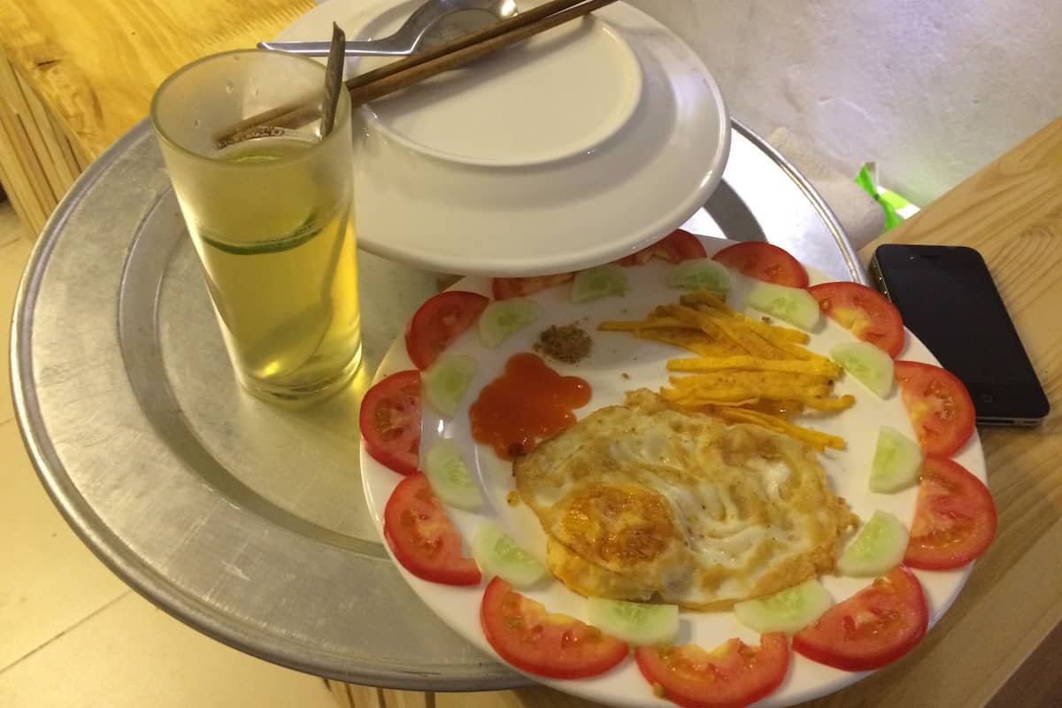 A plate of food with a fried egg, tomato slices, and cucumber, accompanied by a glass of yellow beverage and chopsticks on a tray.