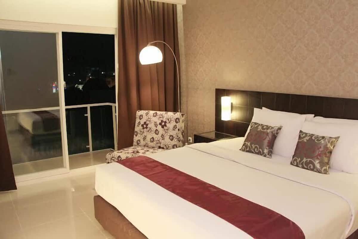 A hotel room with a large bed, a nightstand, a lamp, a chair, and a view of the city through a large window.