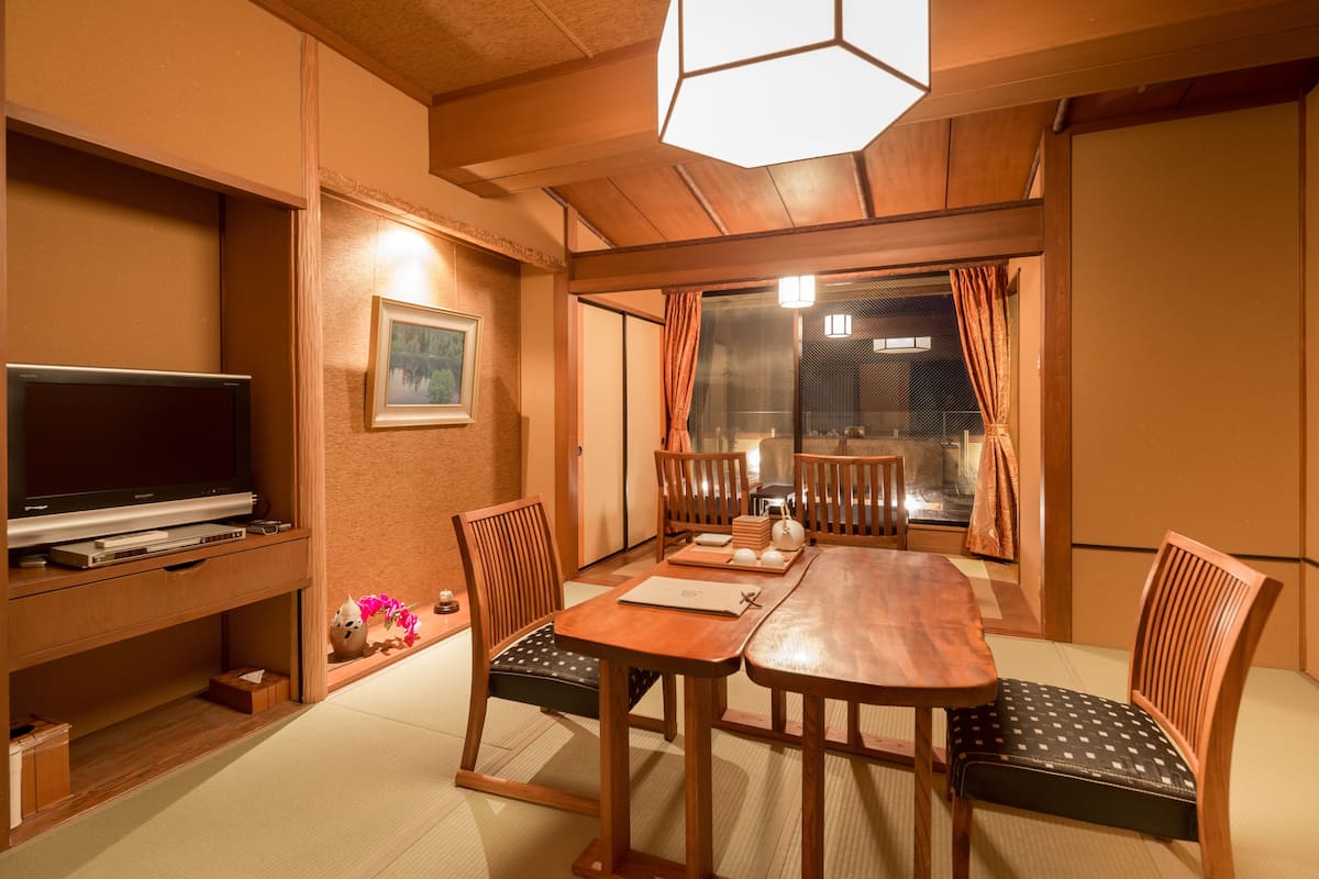 A traditional Japanese-style room with tatami flooring, a low table with cushions, a television on a wooden stand, and sliding doors leading to a balcony.