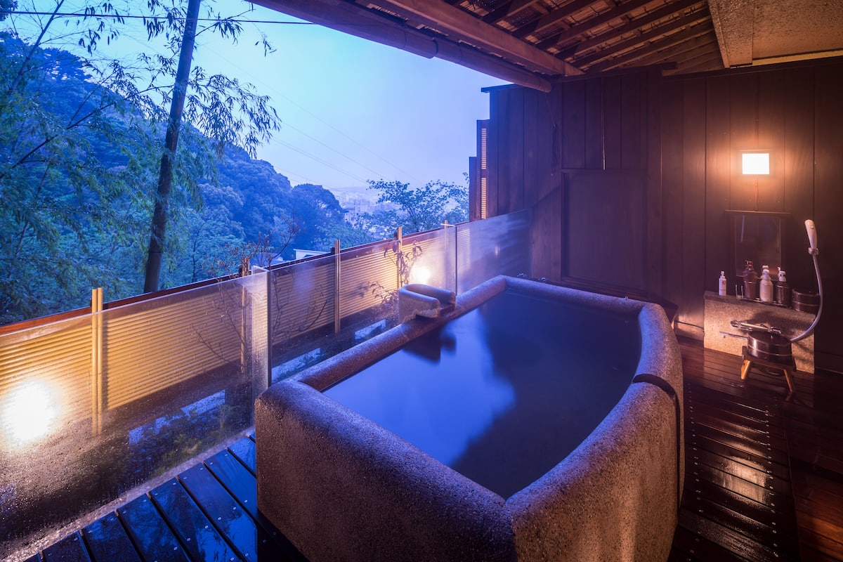 A hot spring bath area with a wooden deck, a stone bath, and a view of trees and hills.