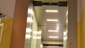 An elevator with a metallic finish and glass panels, equipped with overhead lighting. - Mosel Hotel Frankfurt (Frankfurt)