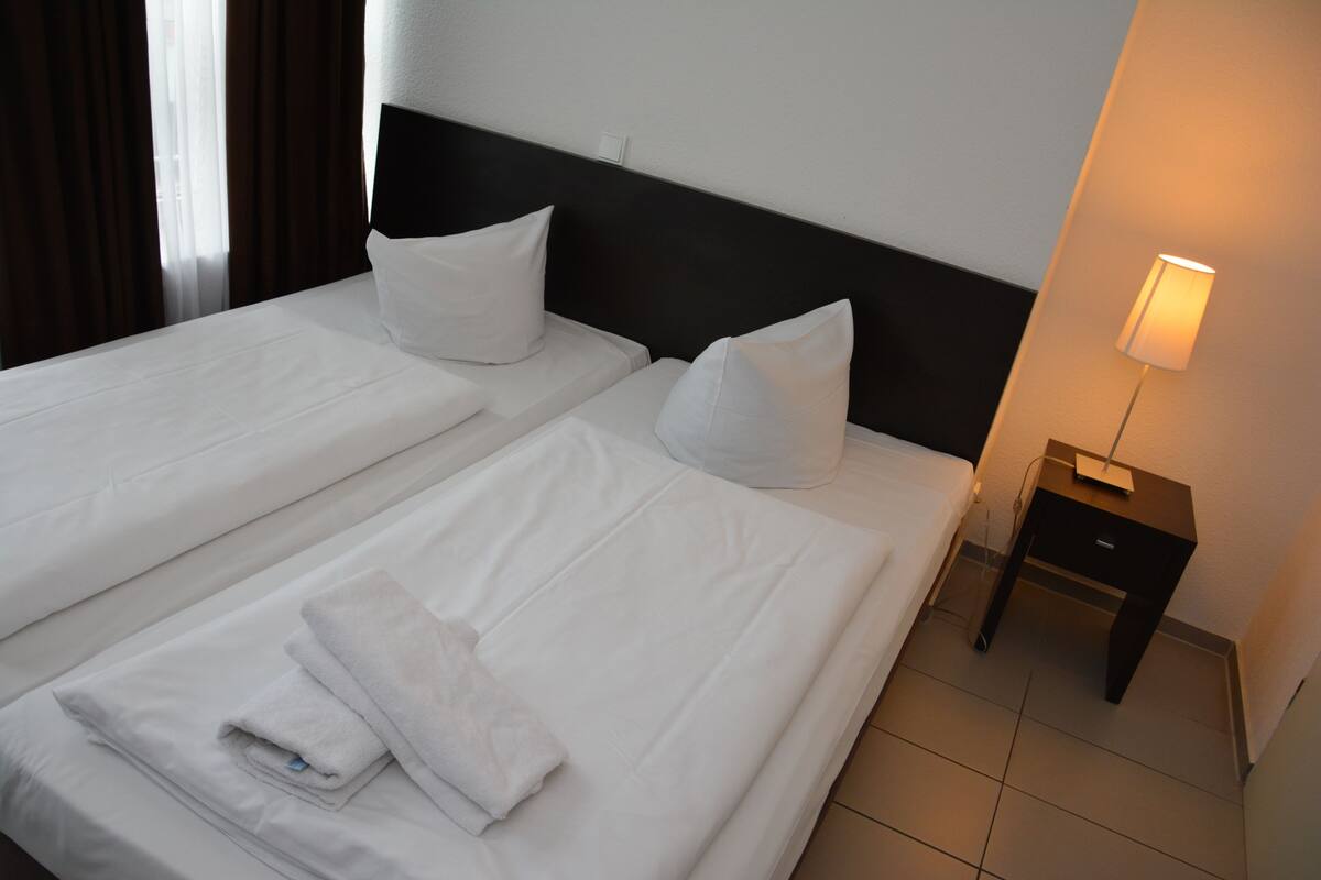 Hotellihuone, jossa on kaksi erillistä sänkyä, molemmissa valkoiset lakanat ja tyynyt, pieni yöpöytä lampun kanssa ja pelkistetty seinä.