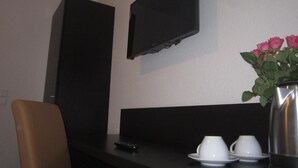 A hotel room with a TV mounted on the wall, a dark wooden desk, a chair, and a vase with pink flowers. - Mosel Hotel Frankfurt (Frankfurt)