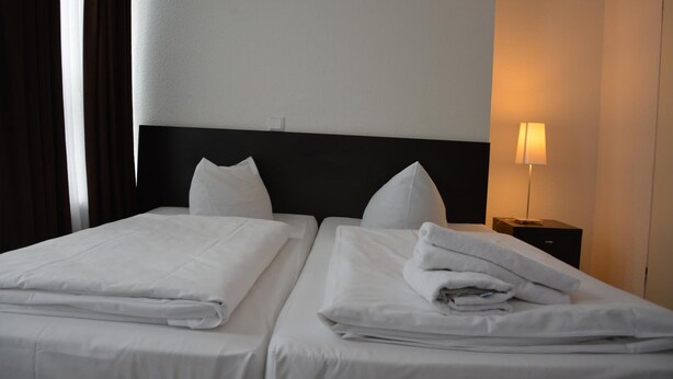 A hotel room with two beds, white linens, a black headboard, a nightstand with a lamp, and a window with curtains. - Mosel Hotel Frankfurt (Frankfurt)