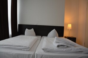 Habitación de hotel con dos camas, ropa de cama blanca, cabecera negra, mesita de noche con lámpara y ventana con cortinas.