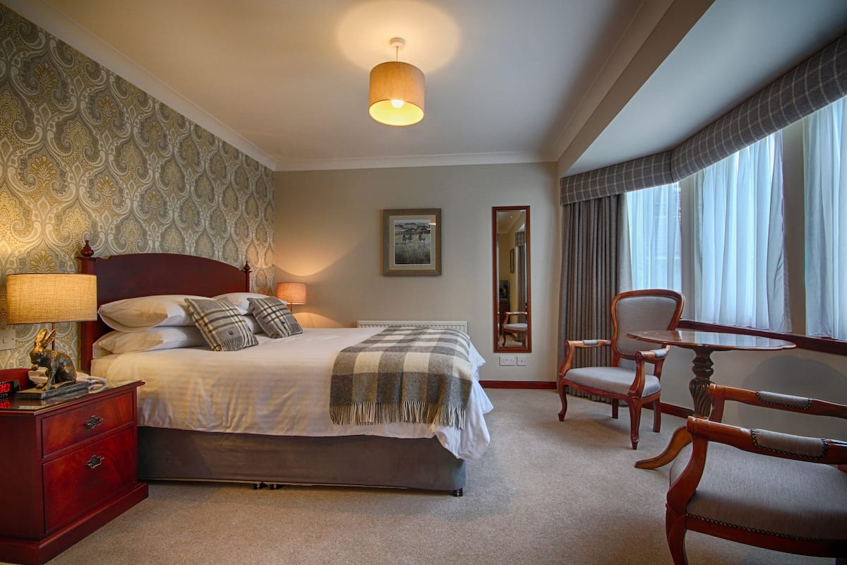 A hotel room with a large bed, a nightstand, a desk, and a chair. There is a window with curtains and a wall with a patterned wallpaper.
