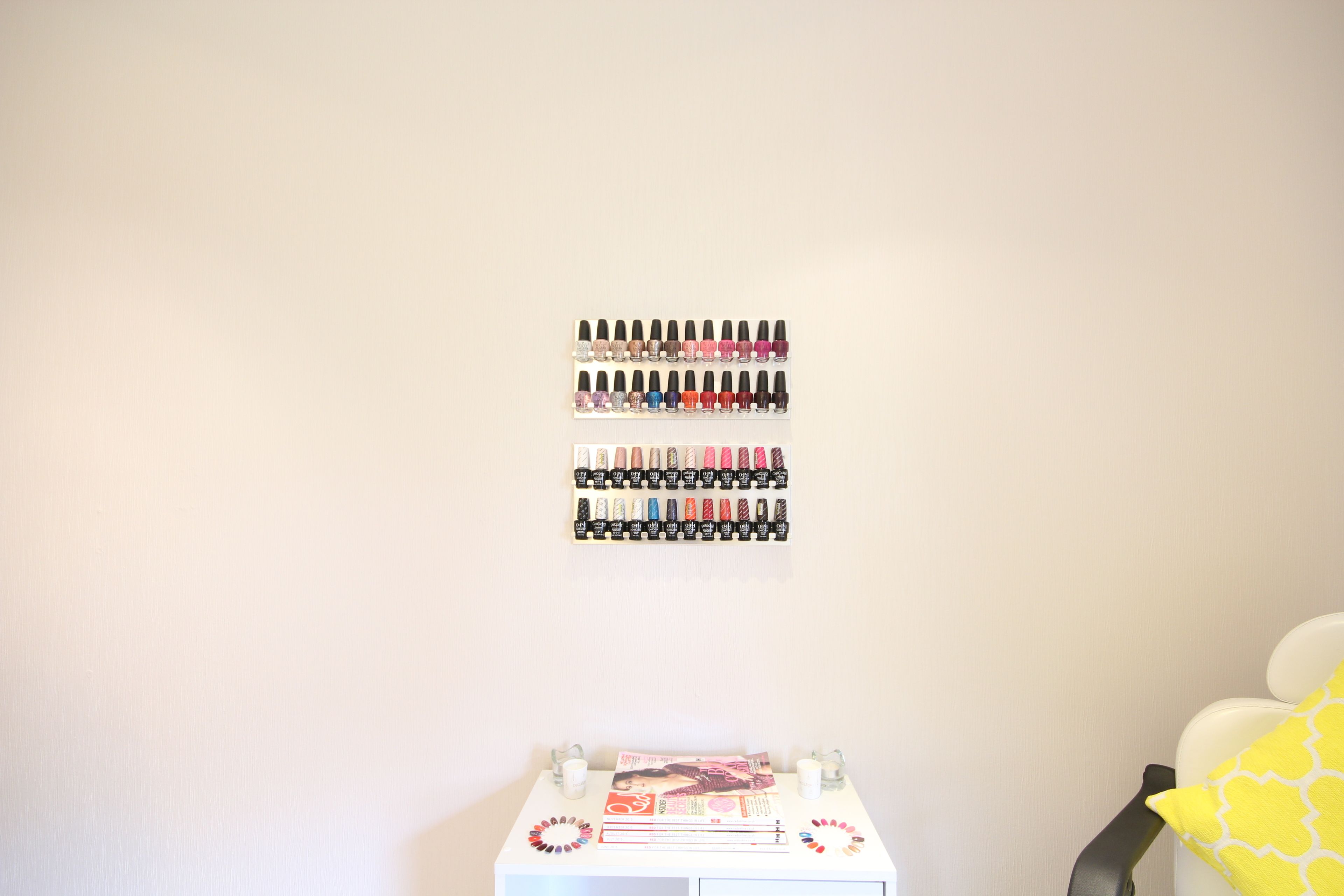 Una exhibición ordenada de esmaltes de uñas en una pared, una mesita con revistas y una taza, y una silla con un cojín amarillo.