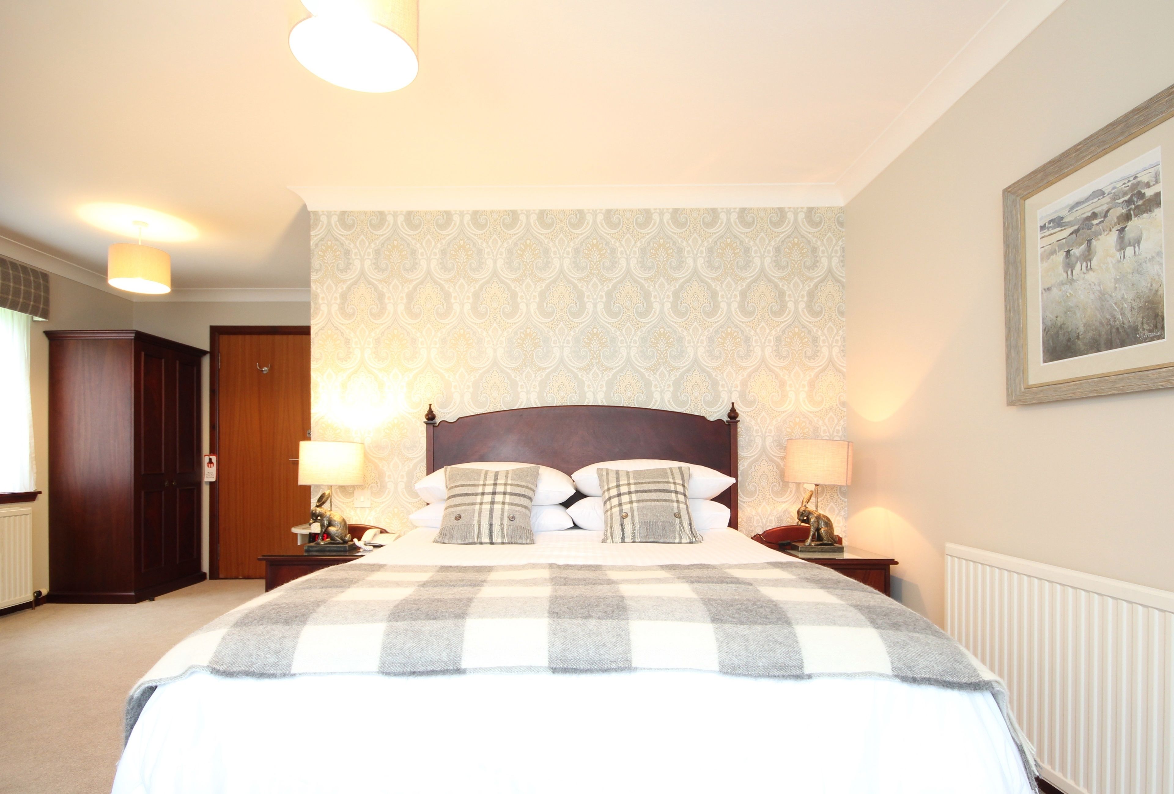 Een netjes ingerichte slaapkamer met een groot bed, twee nachtkastjes met lampen, een houten garderobe en behang met een patroon.