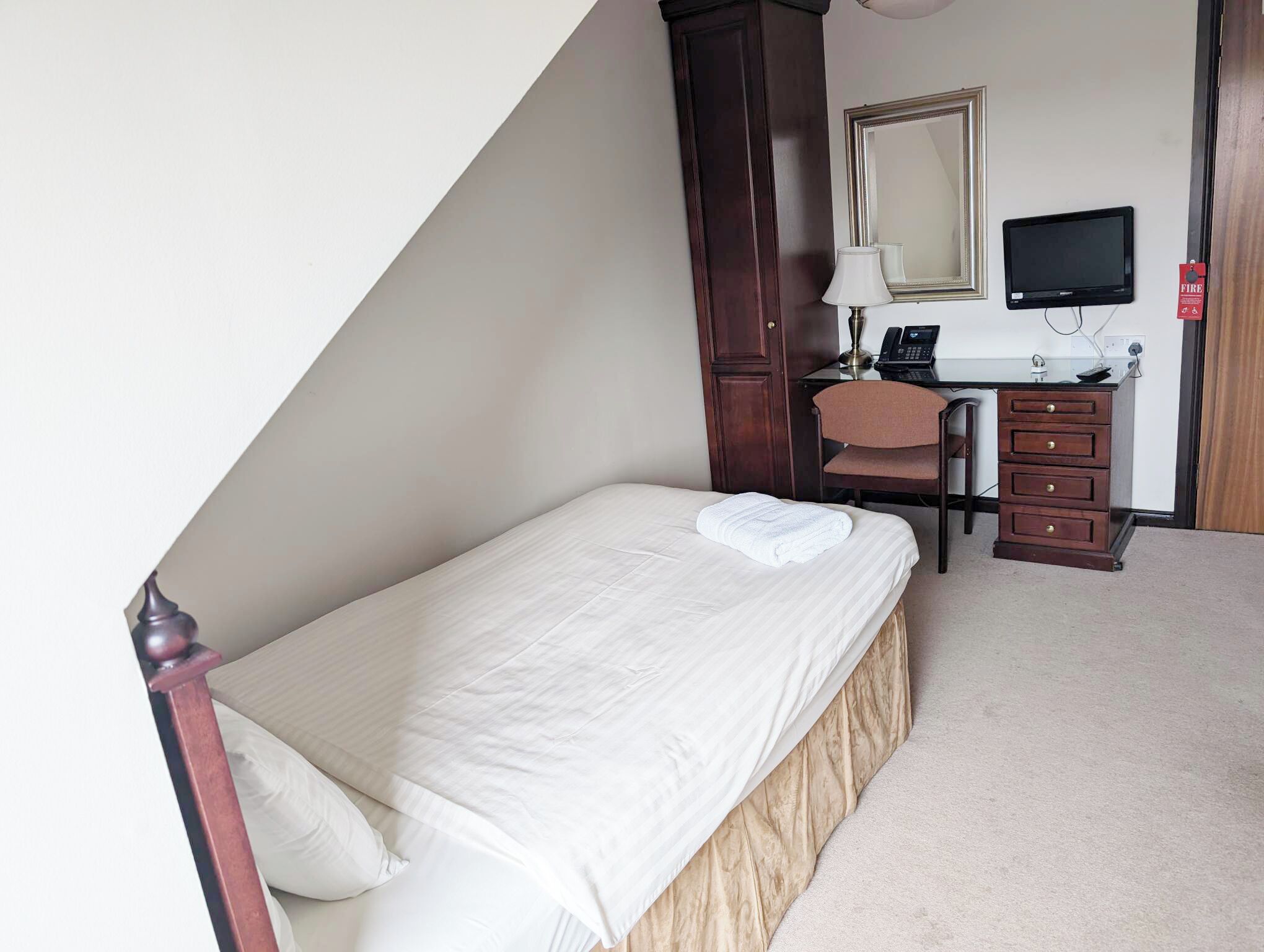 A single bed with a white comforter and a folded towel, a desk with a computer and a telephone, a chair, and a mirror on the wall.