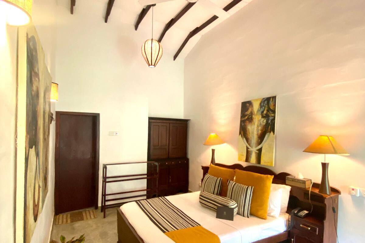 A bedroom with a bed, bedside lamps, a nightstand, a painting on the wall, a door, and a potted plant.