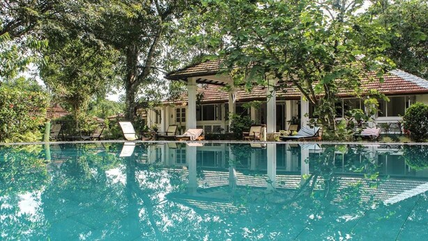 A pool area with lounge chairs and a pergola, surrounded by lush greenery and a building with a tiled roof. - Plantation Villa Ayurveda Yoga Resort (Dodangoda)