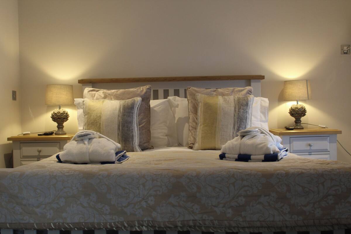 Cama bem arrumada com colcha estampada, dois travesseiros e duas toalhas dobradas na cabeceira. Duas mesas de cabeceira com luminárias e objetos decorativos.