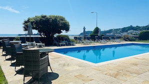 A swimming pool with lounge chairs and umbrellas, surrounded by stone tiles and a view of the sea. - La Haule Manor (St. Brelade)