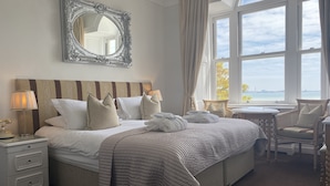 A hotel room with a large bed, a bedside table with a lamp, a chair, and a view of the sea through large windows. - La Haule Manor (St. Brelade)