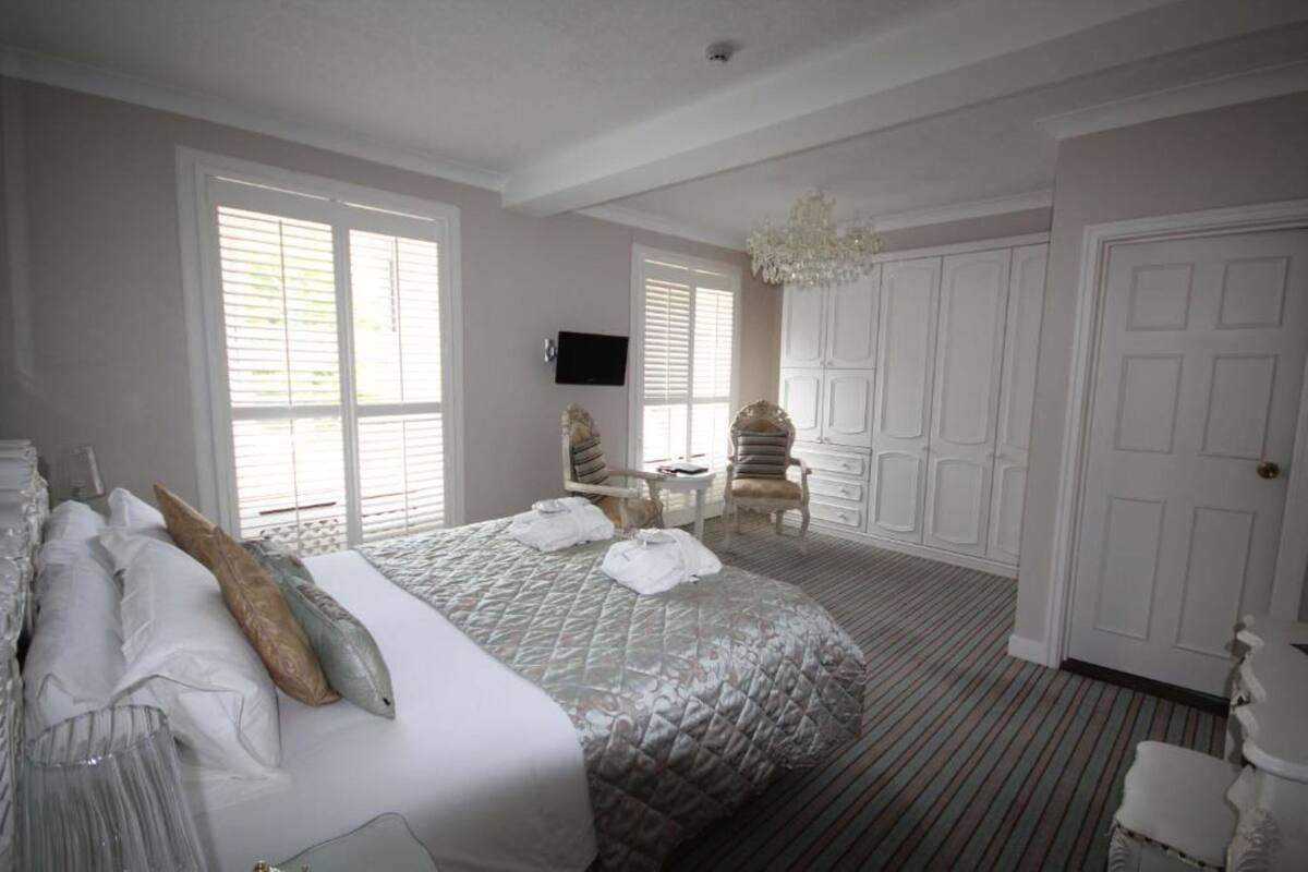 Quarto de hotel com uma cama grande, duas cadeiras, um candelabro, uma televisão e um guarda-roupa.