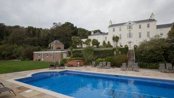 Une piscine avec des chaises longues, un bâtiment en pierre et une grande maison blanche dotée d’un jardin.