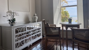 A room with a white console table, two wicker chairs, a small round table with a potted plant, and a view of the ocean through a window with white shutters. - La Haule Manor (St. Brelade)